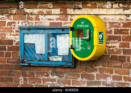 La vita di emergenza salvataggio defibrillatore & vecchio villaggio bacheca affiancati montati su esterno muro di mattoni - Burnby Village, nello Yorkshire, Inghilterra, Regno Unito. Foto Stock