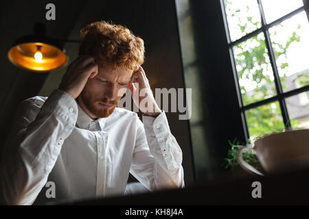 Close-up ritratto di Giovane readhead barbuto oberati di lavoro Uomo in camicia bianca di toccare la sua testa mentre è seduto in ufficio Foto Stock