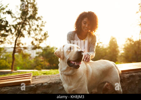 I giovani africani negligente signora seduta sul banco in posizione di parcheggio e abbracciando il suo cordiale cane labrador Foto Stock