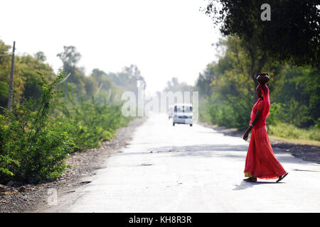 Donna indiana in rosso vestito tradizionale che trasportano una pentola sul suo capo e attraversando la strada in Rajasthan, India. Foto Stock