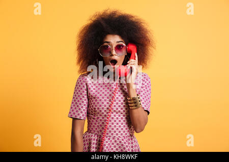 Close-up foto di sorpresa retrò donna con acconciatura afro azienda telefono retrò, guardando la telecamera, isolate su sfondo giallo Foto Stock