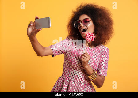 Ritratto di una bella risata afro donna americana in stile retrò vestiti holding lollipop stando in piedi e tenendo un selfie isolate su fondo giallo Foto Stock