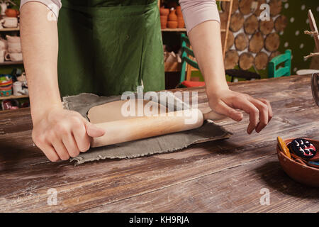 Close-up di una donna potter in un grembiule verde lanciando un rotolamento argilla marrone con un mattarello su un tessuto speciale su un tavolo di legno per fare una piastra Foto Stock