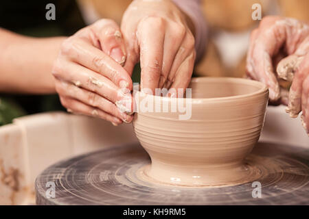 Un close-up di una donna potter insegna una donna come stampare correttamente una ciotola di argilla marrone su un tornio del vasaio in una luminosa officina, vista laterale o di formazione Foto Stock