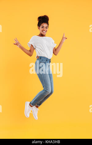 Foto di divertenti african gioiosa signora in bianco maglia e jeans jumping e sorridente isolato Foto Stock