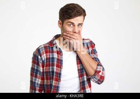 Ritratto di una sorpresa giovane uomo che copre la bocca con la mano e guardando la telecamera isolate su sfondo bianco Foto Stock