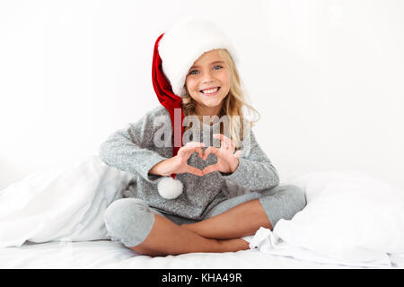 Graziosa bambina Santa's hat che mostra segni di cuore durante la seduta con le gambe incrociate sul letto bianco Foto Stock