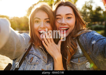 Due giovani allegri brunette donna in jeans usura avendo divertimento tenendo selfie sul telefono cellulare, per esterno Foto Stock