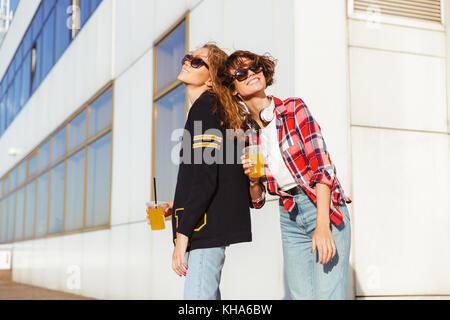 Due allegri ragazze adolescenti in occhiali da sole di bere succo di arancia permanente, mentre all'aperto Foto Stock