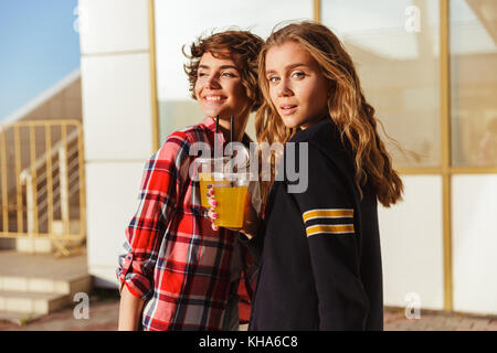 Due allegri giovani ragazze adolescenti in occhiali da sole di bere succo di arancia con una cannuccia mentre in piedi e guardando la fotocamera all'aperto Foto Stock