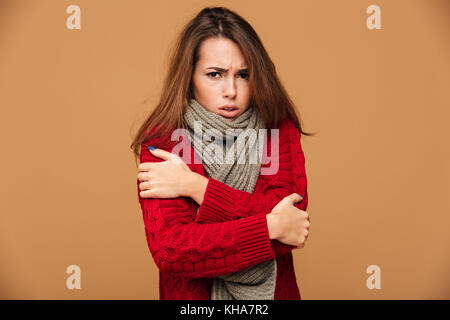 Ritratto di congelamento triste brunette woman in red felpa lavorata a maglia brividi mentre abbraccio di sé e guardando la telecamera, isolato su sfondo beige Foto Stock