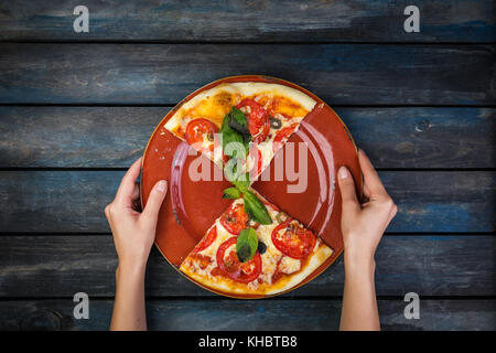 Donna di mani tenendo una piastra con due pezzi di pizza margherita con le fette di pomodoro, le olive e le foglie di basilico. su legno scuro dello sfondo. vista dall'alto. Foto Stock