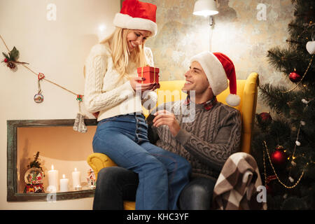 Felice uscito dalla donna bionda in Santa's hat azienda confezione regalo mentre è seduto in poltrona con il marito Foto Stock