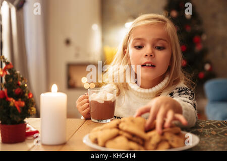 Close-up foto di felice bambina in maglia maglione holding tazza di cioccolata calda, tenendo cookie sulla mattina di Natale Foto Stock