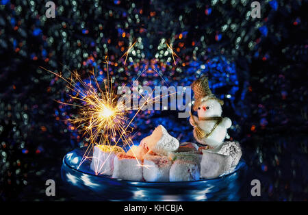 Poco pupazzo di neve con fuochi d'artificio brilla su di una piastra con marshmallows e dolci orientali. Il telaio orizzontale. Foto Stock