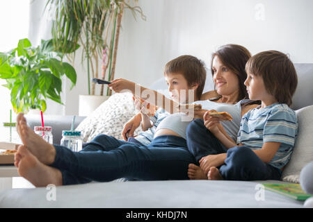 Felice famiglia giovane, donna incinta e due ragazzi, mangiare una gustosa pizza a casa seduti sul divano guardando la TV e avente una risata Foto Stock