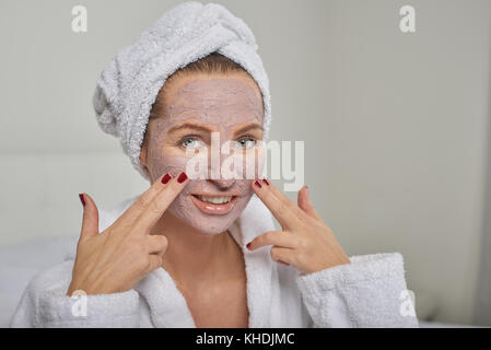 Donna attraente in un bianco accappatoio in spugna applicando una maschera per il viso mentre cercano in un palmare mirror come lei stessa vi vizierà con un trattamento di bellezza Foto Stock