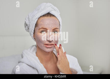 Donna attraente in un bianco accappatoio in spugna applicando una maschera per il viso mentre cercano in un palmare mirror come lei stessa vi vizierà con un trattamento di bellezza Foto Stock
