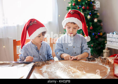 Due simpatici ragazzi con santa hat, preparazione di biscotti a casa, albero di Natale dietro di loro Foto Stock