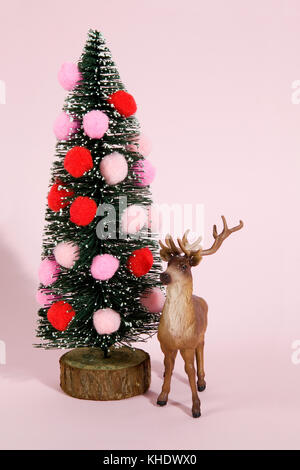 Snowy decorate abeti e albero di Natale decorato con pompon con figurine renne accanto ad esso su un vivace colore rosa sfondo. Il minimo ancora vita Foto Stock