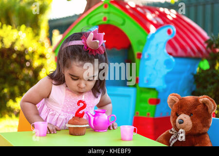 Baby toddler girl in outdoor seconda festa di compleanno soffiando candela su pirottini. orsacchiotto come migliore amico, Playhouse e set per il tè. abito rosa e corona. Foto Stock