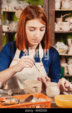 I capelli rossi donna potter scolpisce da una argilla marrone candeliere e colle dettagli convessa di argilla con un pennello e una argilla liquida rende contro il backgr Foto Stock