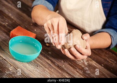 Un close-up di una femmina di potter in una camicia di denim e grembiule detiene una argilla marrone in forma di una sfera, massaggia la creta e rende in esso un foro per effettuare una de Foto Stock