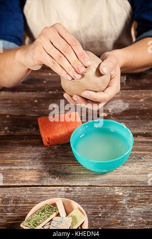 Un close-up di una femmina di potter in jeans e maglietta grembiule è in possesso di una argilla marrone in forma di una sfera, impastare la creta per iniziare a scolpire il vaso Foto Stock