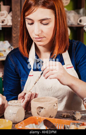 I capelli rossi donna potter scolpisce da una argilla marrone candeliere e colle dettagli convessa di argilla con un pennello e una argilla liquida rende contro il backgr Foto Stock