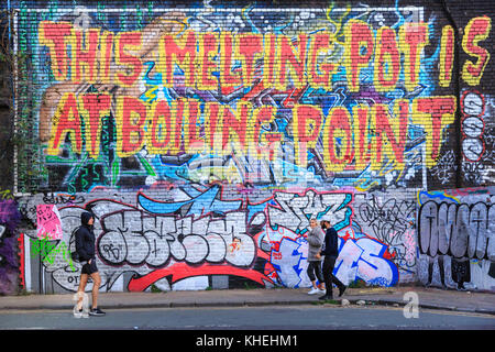 La gente camminare passato grafitti street art sloagan 'questo melting pot è al punto di ebollizione' con commento sociale in Shoreditch, London, Regno Unito Foto Stock