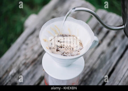 Deliziosi caffè pourover fioritura mentre l'uomo si versa acqua nel gocciolatore a terra i chicchi di caffè. picnic estivo sul vecchio tavolo in legno backgrou Foto Stock