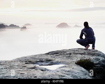 Momento di solitudine su esposta vetta rocciosa. man in black godono di una vista meravigliosa. escursionista sedersi sul picco di roccia e guardare nella nebbia colorata e la nebbia Foto Stock