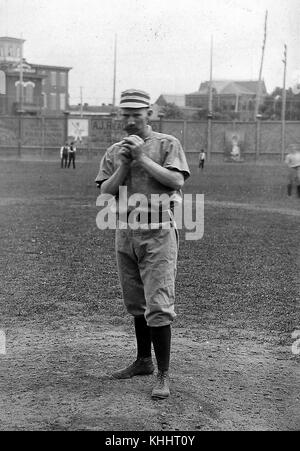 Un ritratto fotografico di Charlie Ferguson in uniforme dei Philadelphia Quakers, nella fotografia che tiene in mano una palla come se si stesse preparando a lanciarla, si trova su un campo da baseball, altri giocatori possono essere visti dietro di lui, oltre gli edifici del campo possono essere visti in città, ha giocato quattro stagioni della Major League Baseball prima di contrarre e morire di febbre tifoide nel 1888, 1885. Dalla New York Public Library. Foto Stock