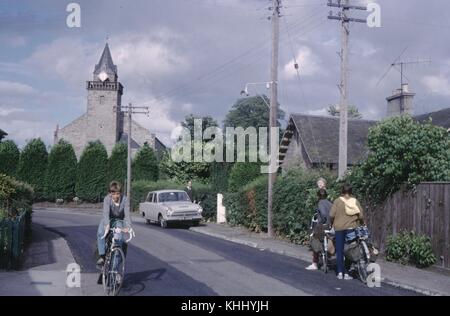 I ciclisti si fermano accanto a una piccola strada locale e guardano una chiesa di pietra mentre un ragazzo passa su una bicicletta nell'altra direzione, Olanda, 1960. Foto Stock