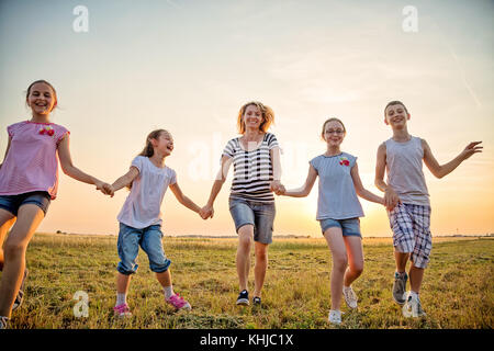 Madre e quattro bambini divertendosi nel campo, tenendo le mani e correre verso la fotocamera all'aperto, in campo estivo al tramonto. che indossa casual c Foto Stock