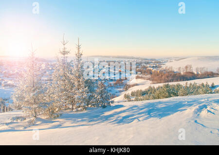 Calda vista illuminata di coperta di neve colline e villaggio con gli abeti in primo piano al tramonto in piccole città europea Foto Stock