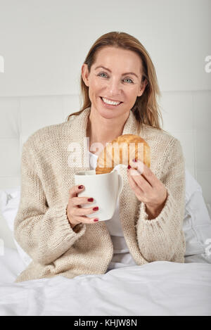 Ritratto di una bella donna sorridente pur avendo la colazione a letto con una tazza di caffè o di latte e un delizioso croissant francesi Foto Stock