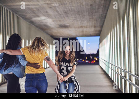 Diversi gruppi di giovani amici di ridere e camminare mano nella mano insieme verso il basso una passerella nella città di notte Foto Stock