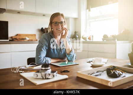 Femmina giovane imprenditore sorridente con fiducia mentre è seduto da solo al suo tavolo da cucina in casa a lavorare sul suo small business Foto Stock