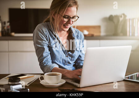 Sorridente giovane donna che lavorano per la sua piccola impresa con un computer portatile mentre è seduto al suo tavolo da cucina a casa Foto Stock