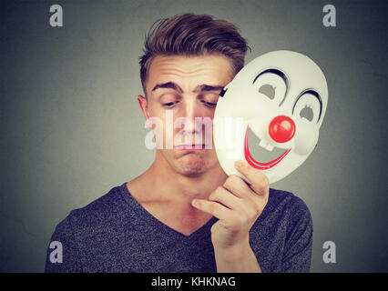 Giovane uomo triste decollare happy clown maschera isolata su sfondo grigio. le emozioni umane. Foto Stock