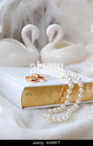 Due fedi nuziali d'oro sulla Bibbia bianca. Perle bianche e due cigni bianchi in porcellana. Sfondo bianco. Versione verticale della foto. Foto Stock