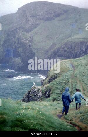 Una fotografia di due escursionisti a piedi lungo un bagno stretto nella parte superiore di un'erba coperto cliff, un'altra persona può essere visto seduto su una roccia che affiora in superficie prima degli altri due escursionisti, le onde dell'oceano può essere visto schiantarsi contro il fondo della scogliera, 1965. Foto Stock