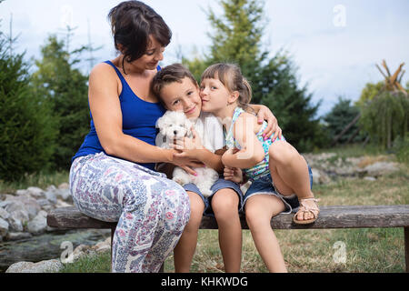 Giovane europeo orientale la madre ed i suoi due piccoli bambini seduti su una panca di legno in una natura sul giorno di estate, coccole e baciare Foto Stock