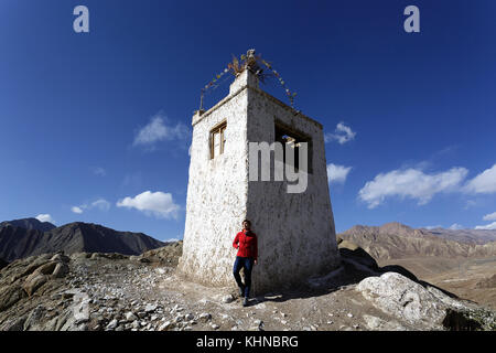 Woman in Red jacket in piedi sul bordo di un tradizionale edificio Ladakhi con bandiere di preghiera, Alchi, Ladakh, Jammu e Kasmhir; India. Foto Stock