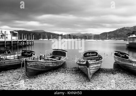 Un bianco e nero foto di imbarcazioni a remi sulla riva di Windermere Near Ambleside con yacht e Lake District colline in lontananza. Foto Stock