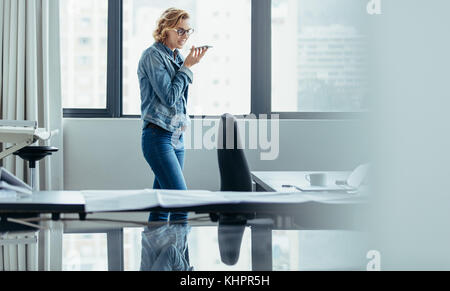 Inquadratura orizzontale di imprenditrice tramite cellulare in ufficio. Architetto femmina parlando al telefono con altoparlante. Foto Stock