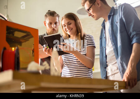 Ritratto di attraente giovane designer che mostra i risultati del lavoro ai suoi colleghi di sesso maschile con l aiuto della tavoletta digitale mentre si lavora insieme su ambizioni Foto Stock