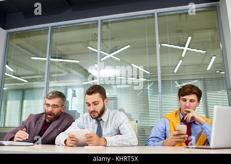 Tre annoiato la gente di affari in attesa di incontro e l'utilizzo dei gadget seduto in fila al tavolo nella sala riunioni Foto Stock