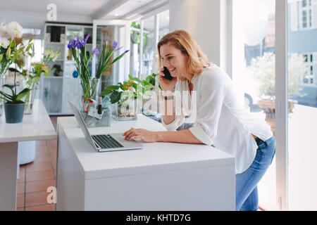 Fiorista femmina parlando al telefono cellulare durante l'utilizzo di laptop in negozio di fiori. Felice giovane donna facendo ordine in vivaio. Foto Stock